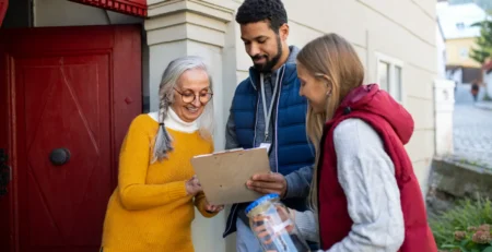 Two volunteers fundraising outside a home as a smiling woman signs a clipboard on her doorstep.